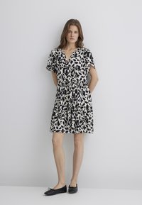 Kurzärmliges Kleid in Creme mit einem schwarzen Blattmuster, mit V-Ausschnitt und gerafftem Bund, kombiniert mit schwarzen Ballerinas.