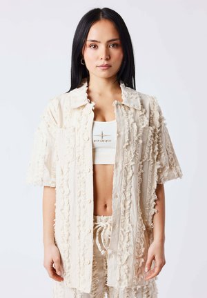 Donna con capelli lunghi neri che indossa una camicia oversize color crema con texture, pantaloncini abbinati e un crop top bianco con logo, in piedi davanti a uno sfondo semplice.