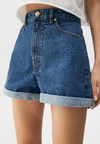 Shorts de mezclilla con cintura alta, dobladillos enrollados, lavado oscuro y costura anaranjada. Presenta dos bolsillos delanteros y un botón metálico estándar.