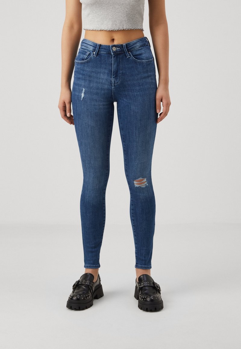 Jean bleu slim avec des détails déchirés sur la cuisse et le genou, porté avec un haut gris court et des mocassins noirs cloutés.
