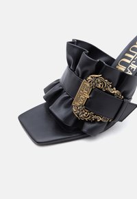 Μαύρο mule από δέρμα με ραμμένη φάσα με βολάν και διακοσμητική χρυσή αγκράφα χαραγμένη με τη φράση «Versace Jeans Couture», με τετράγωνη σχεδίαση στο μπροστινό μέρος.