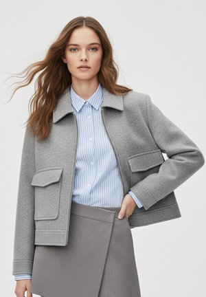 Szara marynarka z mieszanki wełny z kołnierzem, dwoma przednimi kieszeniami i zamkiem błyskawicznym, noszona na jasno-niebiesko-białej koszuli w paski, zestawiona z szarą spódnicą.