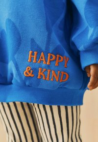 Blauer Sweatshirt mit orangefarbenem gesticktem Text "HAPPY & KIND." Verfügt über gerippte Bündchen und Saum, kombiniert mit schwarz-weißen gestreiften Hosen.