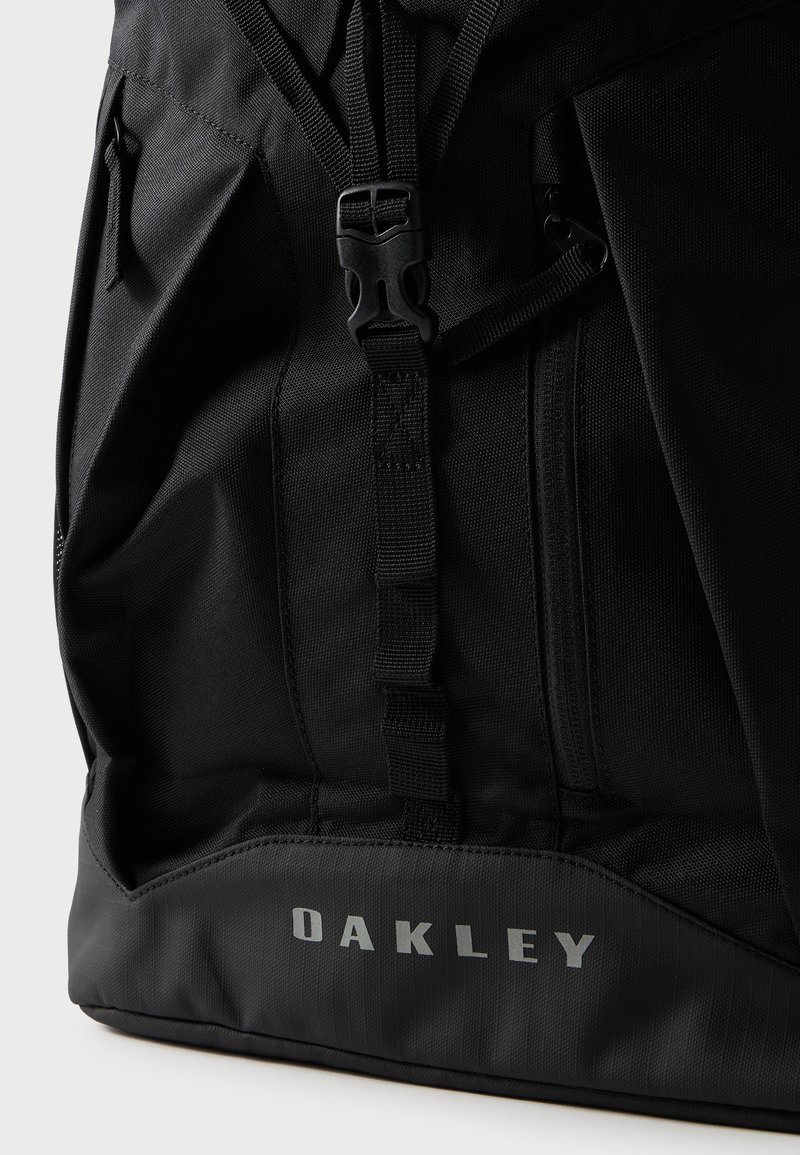 Schwarzer Oakley-Rucksack mit Schnalle am vorderen Gurt und seitlicher Reißverschlusstasche, zeigt strapazierfähige Stofftextur und Markenlogo unten.