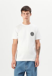 Hvit bomull T-skjorte med et sirkulært svart logo på venstre bryst. Modellen har på seg lyseblå denimbukser, med synlig tatovering på høyre arm.