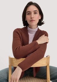 Bordeaux-roter, gerippter Strickcardigan mit einem lockeren Schnitt, ausgestattet mit einem Schalkragen und langen Ärmeln, über einem helllila Rollkragenpullover getragen.