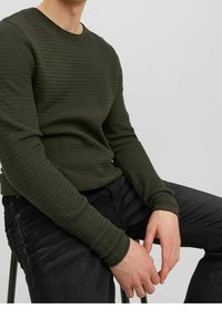Ciemnozielona, teksturowana koszulka z długim rękawem i okrągłym dekoltem; w zestawieniu z czarnymi spodniami. Koszulka ma wzór tkany i dopasowane rękawy.