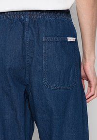 Ciemnoniebieskie dżinsy z tylną kieszenią z białą etykietą z napisem "Calvin Klein". Elastyczny pas i prosty krój nogawek.