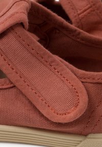 Scarpa in tela color ruggine con una fascia regolabile, tessuto texture e suola flessibile in gomma crema. Sono visibili dettagli cuciti.