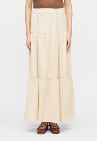 Γυναίκα που φοράει μακρύ, πολυεπίπεδο κρεμ φούστα με ελαστική μέση, καφέ μπλουζάκι και καφέ δερμάτινα παπούτσια με κορδόνια, όρθια μπροστά σε απλό φόντο.