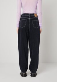 Persona, kas valkā melnas augsta jostas plata izgriezuma džinsas ar redzamām aizmugurējām kabatām un melnas masīvas kurpes, stāv pret vienkrāsainu gaišu fonu.