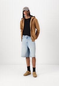 Haut à capuche zippé marron sur un t-shirt noir, shorts en denim bleu et baskets beiges avec des chaussettes noires. Une casquette grise complète la tenue décontractée.