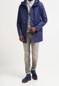 Man in een blauwe regenjas, lichtgrijze trui, gestreept shirt, beige jeans en blauwe suède laarzen, poseert tegen een effen achtergrond.