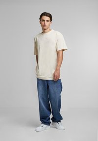 Lysbeige oversized t-shirt med korte ærmer, broderet logo på brystet, parret med løse blå denimjeans og hvide sneakers.