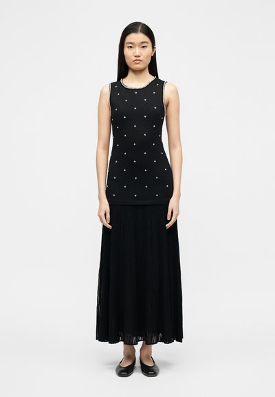 Γυναίκα που στέκεται φορώντας αμάνικο μαύρο τοπ με μικρά μοτίβα σταυρού και μακριά πλισέ μαύρη φούστα με μαύρα επίπεδα παπούτσια.