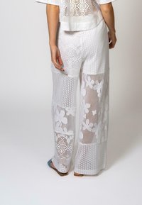 Pantalons en crochet blancs avec un motif ouvert et texturé orné de designs floraux, d'une coupe décontractée. L'ensemble est assorti de sandales plates.