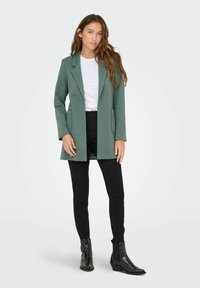 Blazer vert texturé avec poches latérales, porté sur un t-shirt blanc et un jean slim noir, associé à des bottines noires.