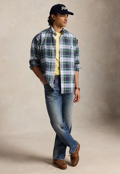 Moški z črno kapo, zeleno-belim karo srajco preko rumene majice, modrimi kavbojkami in rjavimi čevlji stoji z rokami v žepih.