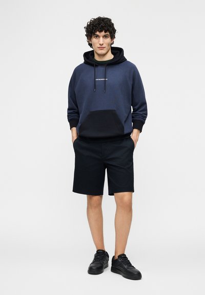 Fiatal férfi áll, sötétkék és fekete kapucnis pulóvert, fekete rövidnadrágot és fekete sportcipőt visel, a kezét zsebre dugva, előre néz egyszerű háttér előtt.