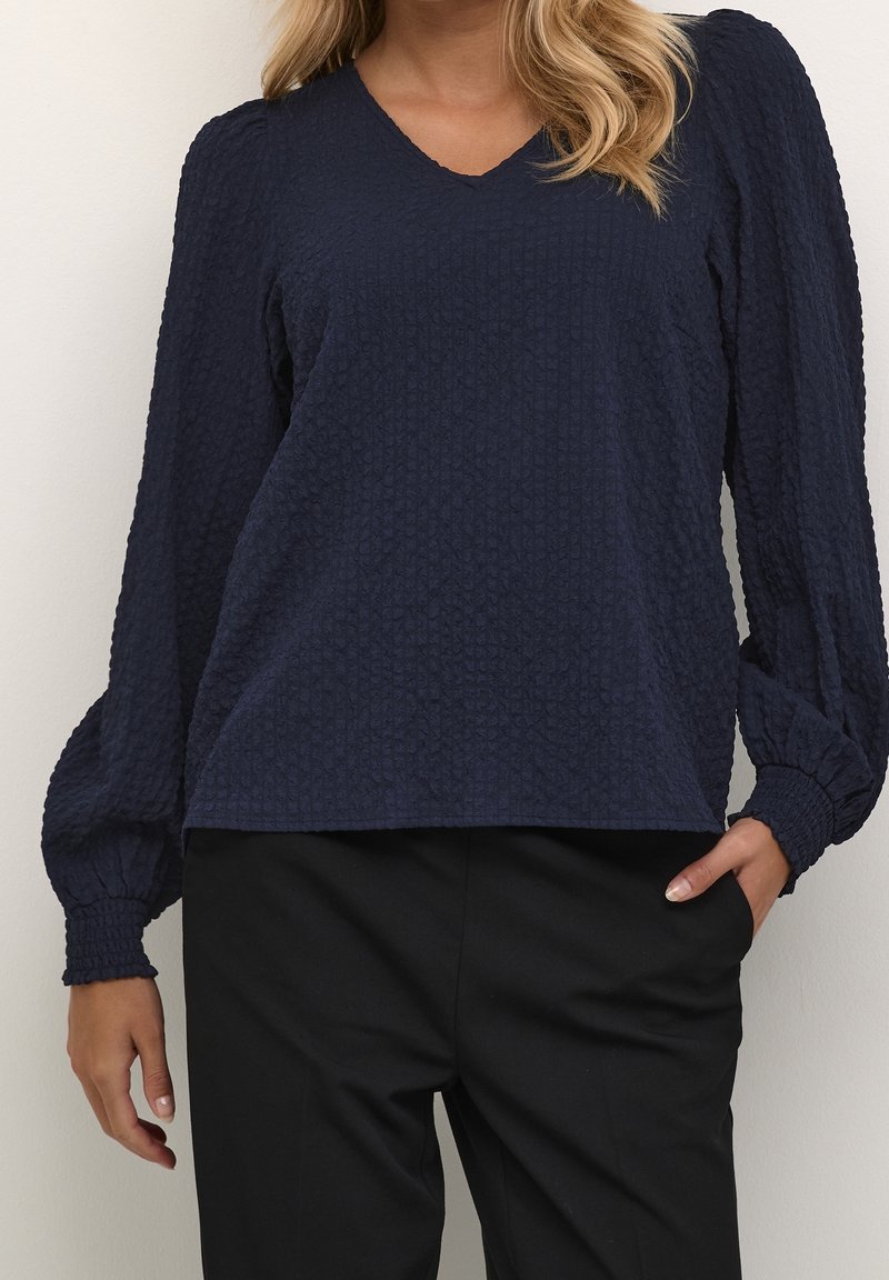 Femme portant un chemisier bleu marine foncé texturé à manches longues bouffantes et un pantalon noir, une main dans la poche et des cheveux blonds visibles.
