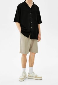 Maglietta nera a maniche corte con bottoni, abbinata a pantaloni corti beige. Sneakers bianche sportive con dettagli color sabbia e calzini bianchi alla caviglia.
