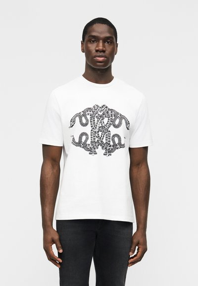 Hombre negro con una camiseta blanca que tiene un diseño abstracto de serpiente en blanco y negro, combinado con vaqueros oscuros, de pie contra un fondo liso.