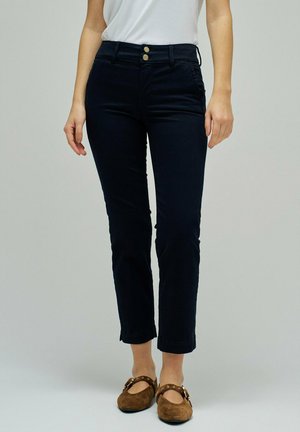 Mujer con pantalones negros recortados con cintura de doble botón y zapatos marrones de ante con hebilla, posando frente a un fondo liso.