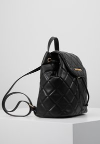 Mustalla tikatulla nahkarepulla, jossa on säädettävät hihnat ja läppäkiinnitys, etupuolella kultasävytteinen logo.