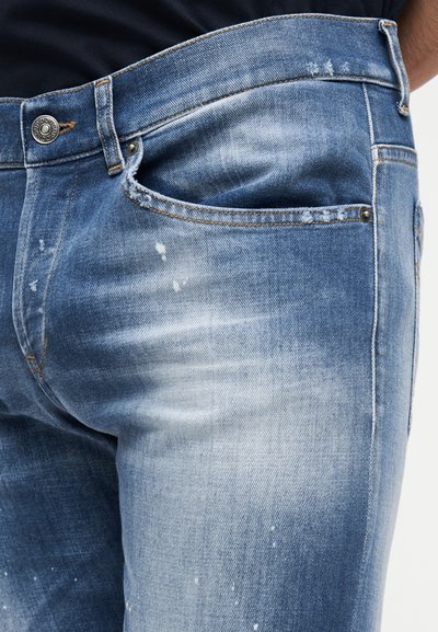 Κοντινή λήψη ενός ατόμου που φοράει ξεθωριασμένα τζιν παντελόνια σε ανοιχτό μπλε χρώμα με μικρές πιτσιλιές μπογιάς και μεταλλικό κουμπί κλεισίματος.