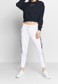 Felpa corta blu navy con logo ricamato, abbinata a pantaloni jogger bianchi con strisce laterali e orli cuffati. Sneakers bianche completano il look.