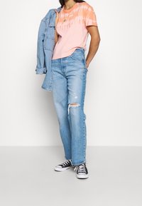 Camiseta de tie-dye rosa claro con acentos naranjas, jeans de mezclilla azul claro desgastados y zapatillas negras de caña baja, terminando con una chaqueta de mezclilla.