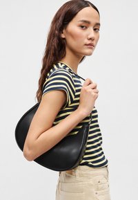 Μαύρη δερμάτινη τσάντα με καμπυλωτό σχήμα, φορεμένη στον ώμο, σε συνδυασμό με κίτρινη και ναυτική μπλούζα με οριζόντιες ρίγες και ανοιχτόχρωμο παντελόνι.