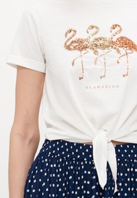 T-shirt blanc à manches roulées, ornée de trois flamants roses en rose doré scintillant et du texte "FLAMAZING." Noué à la taille, associé à une jupe à pois.