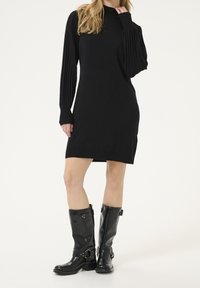 Robe noire avec manches longues plissées, corsage ajusté et ourlet arrivant au genou. Associée à des bottes noires montantes à boucles.
