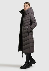 Donna che indossa un lungo cappotto imbottito grigio scuro con un colletto alto, in piedi con le mani in tasca, pantaloni neri e stivali neri.