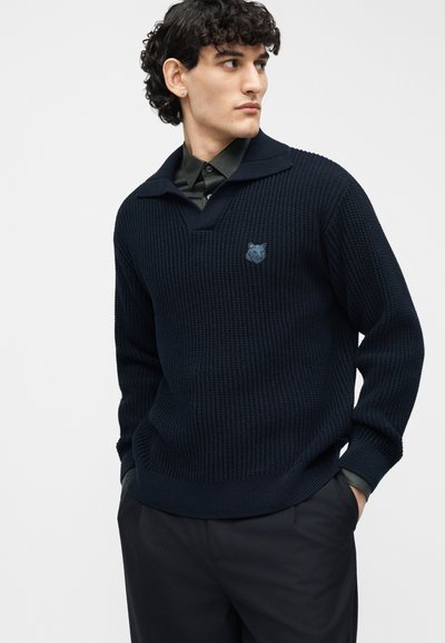 Jeune homme aux cheveux bouclés portant un pull en maille bleu marine foncé par-dessus une chemise noire, les mains dans les poches, regardant vers sa gauche.