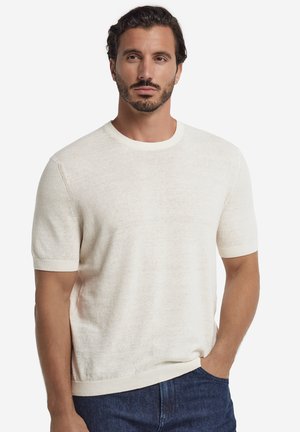 Mand med mørkt hår og skæg iført en lys beige kortærmet sweater og mørkeblå jeans, stående med den ene hånd i lommen.