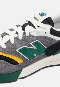 Šedé semišové tenisky s zelenými, žlutými a černými akcenty, s bílou podrážkou a výrazným logem New Balance v kontrastní barvě.