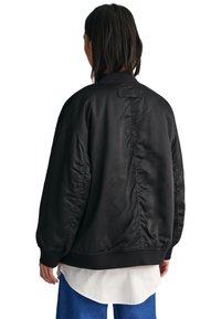 Veste bomber noire avec une finition lisse et brillante, des poignets côtelés et une coupe ample. Présente un design simple et minimaliste.