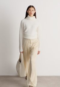 Γυναίκα που περπατά μέσα σε κλειστό χώρο φορώντας λευκό πουλόβερ με ζιβάγκο, μπεζ φαρδιά παντελόνια και κρατώντας μια μεγάλη μπεζ τσάντα ώμου.