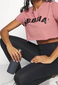 Camiseta corta rosa con texto de logo negro, combinada con leggings negros. Persona sostiene una botella de agua metálica gris, sentada sobre una superficie clara.