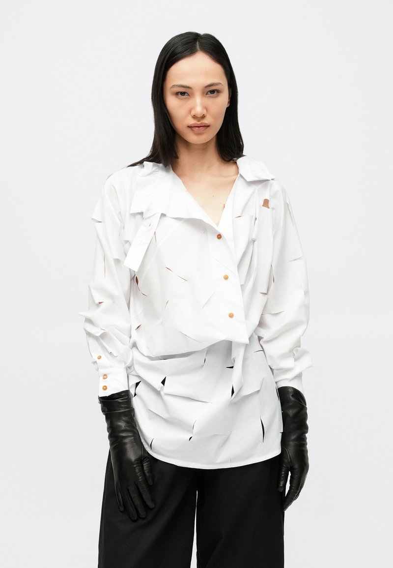 Λευκό πουκάμισο με μακριά μανίκια και λεπτομέρειες κοψίματος, κλειστό με κουμπιά στο μπροστινό μέρος και κλασικό γιακά, συνδυασμένο με μαύρα δερμάτινα γάντια και φαρδιά μαύρη παντελόνα.