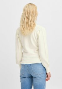 Krämfärgad stickad tröja med puffärmar, ribbad fåll och mönstrad detalj vid midjan, parat med ljusblå jeans.