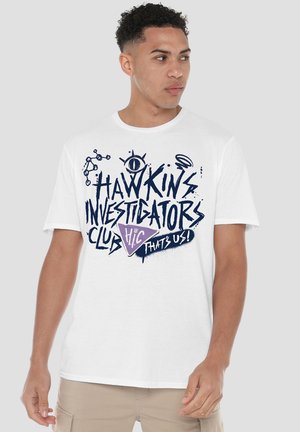 Young man wearing white T-shirt with navy text "Hawkins Investigators Club" and purple triangle logo, beige pants, looking right.