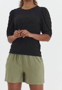 Kvinna i en strukturerad svart topp med korta ärmar och puffiga axlar, iförd ljusgröna shorts med dragsko, stående med händerna ihopknäppta.