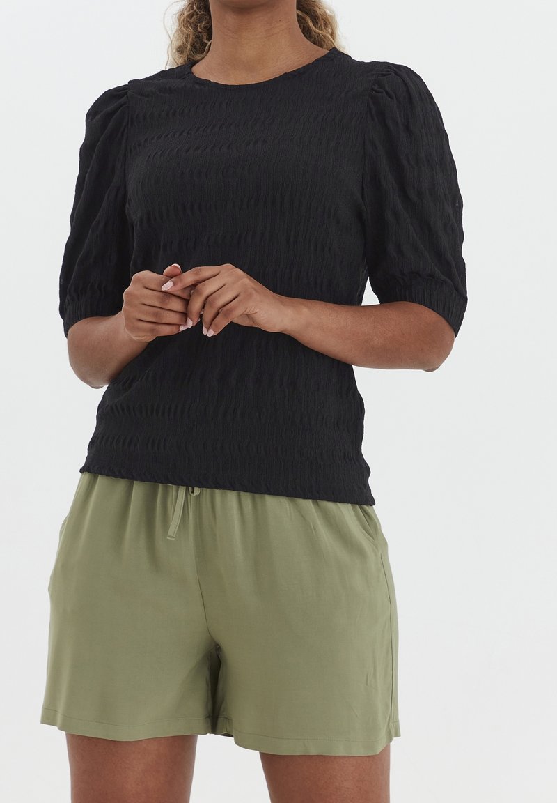 Kvinna i en strukturerad svart topp med korta ärmar och puffiga axlar, iförd ljusgröna shorts med dragsko, stående med händerna ihopknäppta.