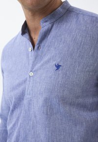 Camisa azul claro, de botones, con cuello mao, confeccionada en tejido texturizado. Presenta un pequeño logotipo de un pájaro bordado en el pecho.
