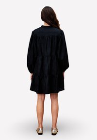 Femme aux longs cheveux bruns portant une robe noire ample à manches bouffantes et des chaussures plates dorées, se tenant de dos contre un fond uni.