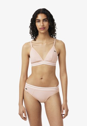Frau trägt hellrosa geripptes Bralette und passende Slips mit Logo-Patches auf der linken Brust und der linken Hüfte, steht vor weißem Hintergrund.