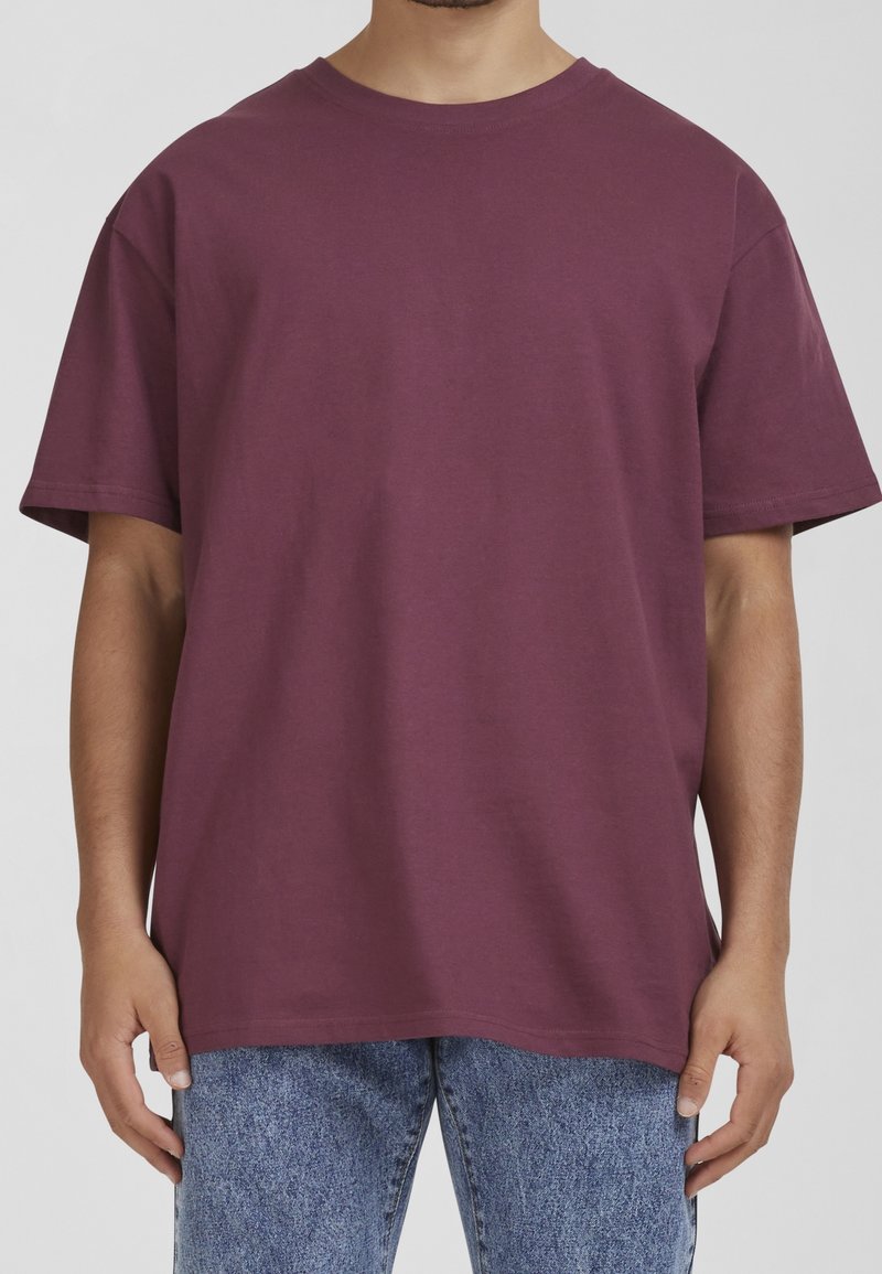 Personne portant un t-shirt uni bordeaux à manches courtes et un jean bleu délavé à l'acide, debout, les mains détendues sur les côtés.